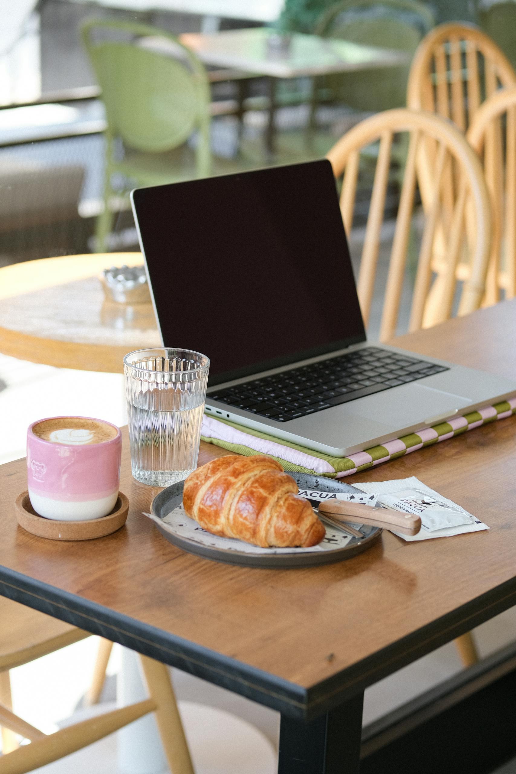 A captivating morning workspace setup in a café with a laptop, coffee, and croissant.